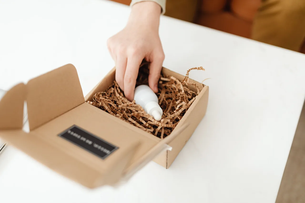 Tay lấy một chai mẫu màu trắng khỏi hộp carton có giấy chèn bảo vệ